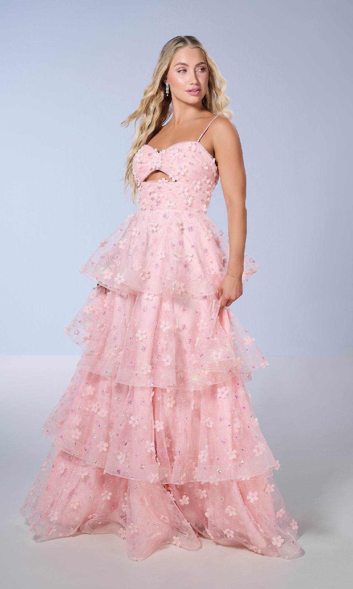A woman stands against a light blue background wearing the Long Prom Dress: Tiffany 16144—a long, tiered pink gown with a ruffled A-line skirt adorned with delicate 3D flowers.